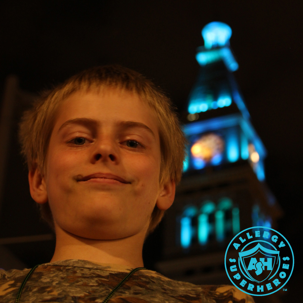 Denver's D&F Clock Tower on 16th Street Mall, with the top floors lit up Teal for Food Allergy Awareness, with a child standing in the foreground | by Food Allergy Superheroes