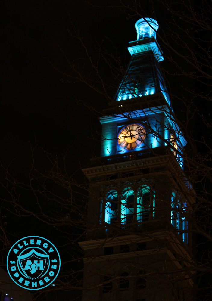 Denver's D&F Clock Tower on 16th Street Mall, with the top floors lit up Teal for Food Allergy Awareness, viewed dramatically through the branches of a tree | by Food Allergy Superheroes