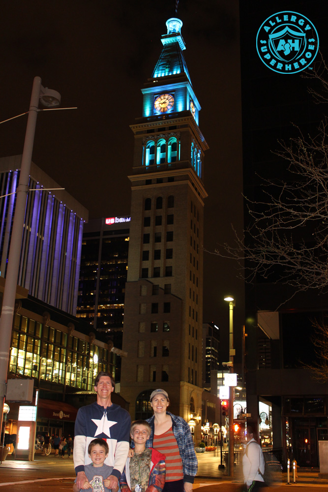 Denver's D&F Clock Tower on 16th Street Mall, with the top floors lit up Teal for Food Allergy Awareness, with the Allergy Superheroes Family standing in front of it | by Food Allergy Superheroes