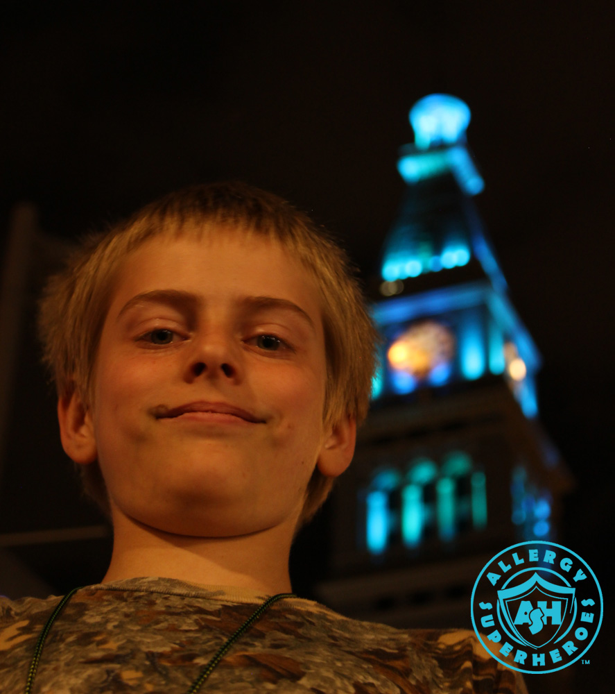 Denver's D&F Clock Tower on 16th Street Mall, with the top floors lit up Teal for Food Allergy Awareness, with a child standing in the foreground | by Food Allergy Superheroes